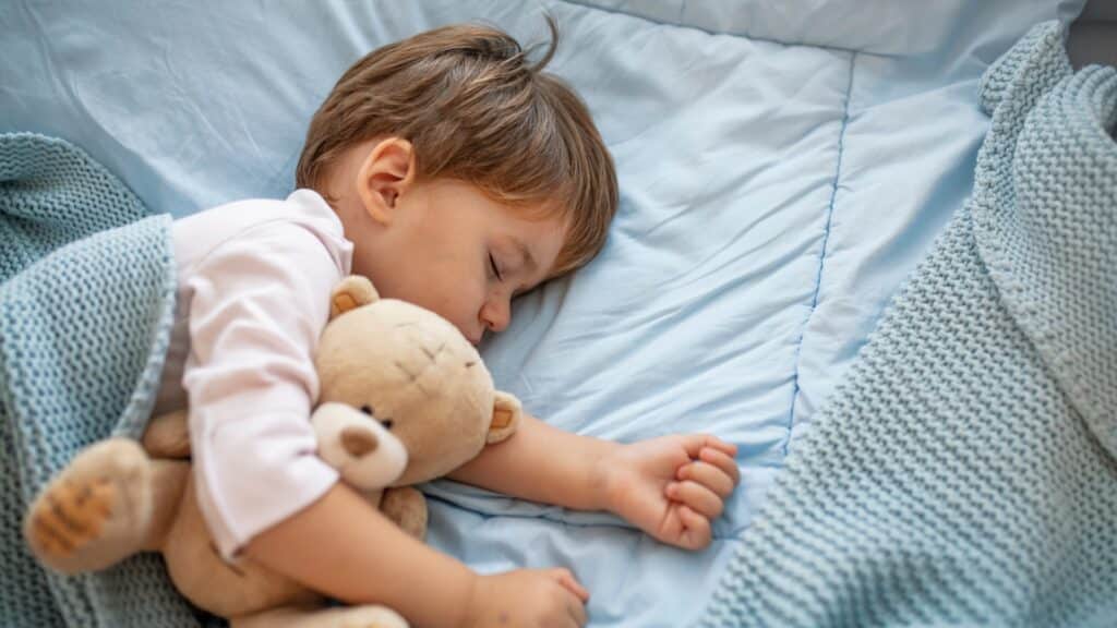 Calm sleeping toddler boy in blue bed sheets cuddling a brown teddy bear