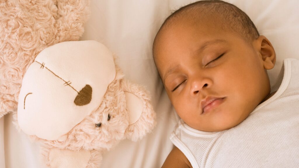 Baby boy asleep on his crib mattress lying next to a fluffy cuddly bear