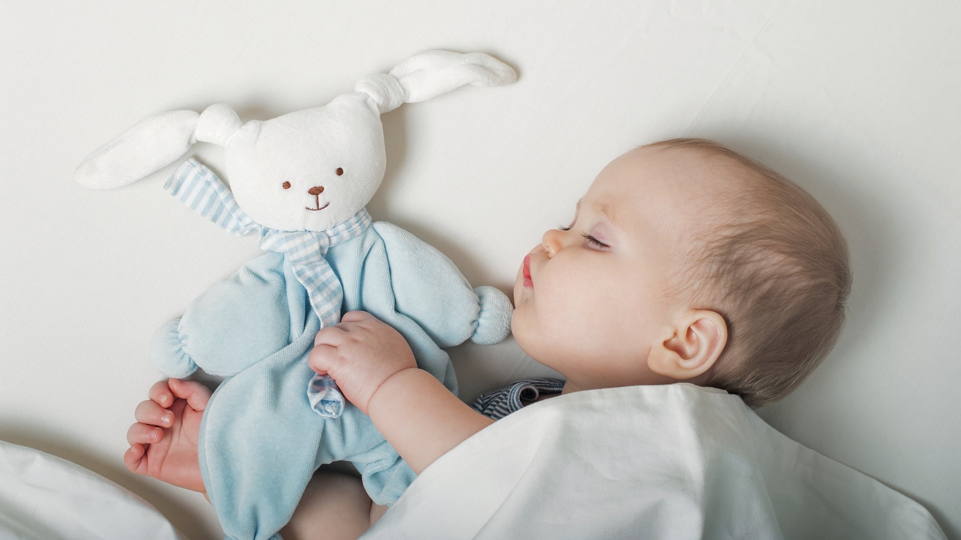 When Can Baby Sleep With Lovey Featured - young baby sleeping next to blue bunny blanket